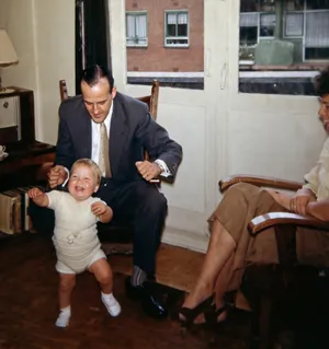 Wim as a toddler in 1960, held upright by the hands by his father in a living room, a woman watching from an armchair